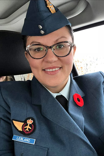 Cheryl LeBlanc smiles into the camera. It is a full frame of Cheryl. She is sitting in a vehicle wearing a blue military uniform with a badge and a name tag on her left lapel and a poppy on her right lapel. She is wearing glasses and a blue peaked military cap.