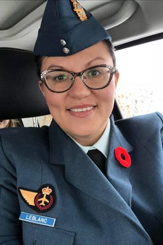 Cheryl LeBlanc smiles into the camera. It is a full frame of Cheryl. She is sitting in a vehicle wearing a blue military uniform with a badge and a name tag on her left lapel and a poppy on her right lapel. She is wearing glasses and a blue peaked military cap.