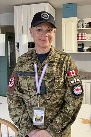 A soldier stands smiling for a photo. She is wearing military fatigues, with a Canadian flag patch on her shoulder. A lanyard with the transgender pride flag colours (light blue, pink and white) hangs around her neck.